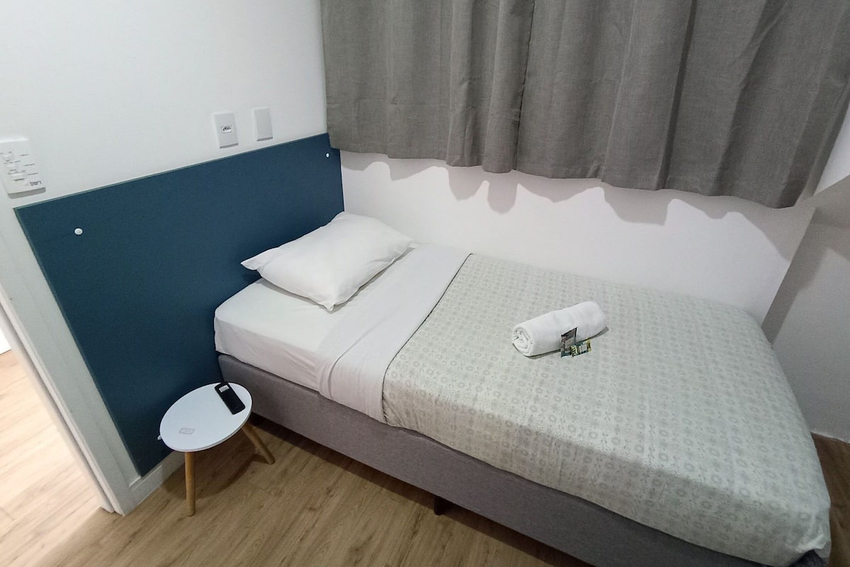 A small, single-bed room with a blue accent wall, a white pillow, a rolled towel, and a small white table with a phone.