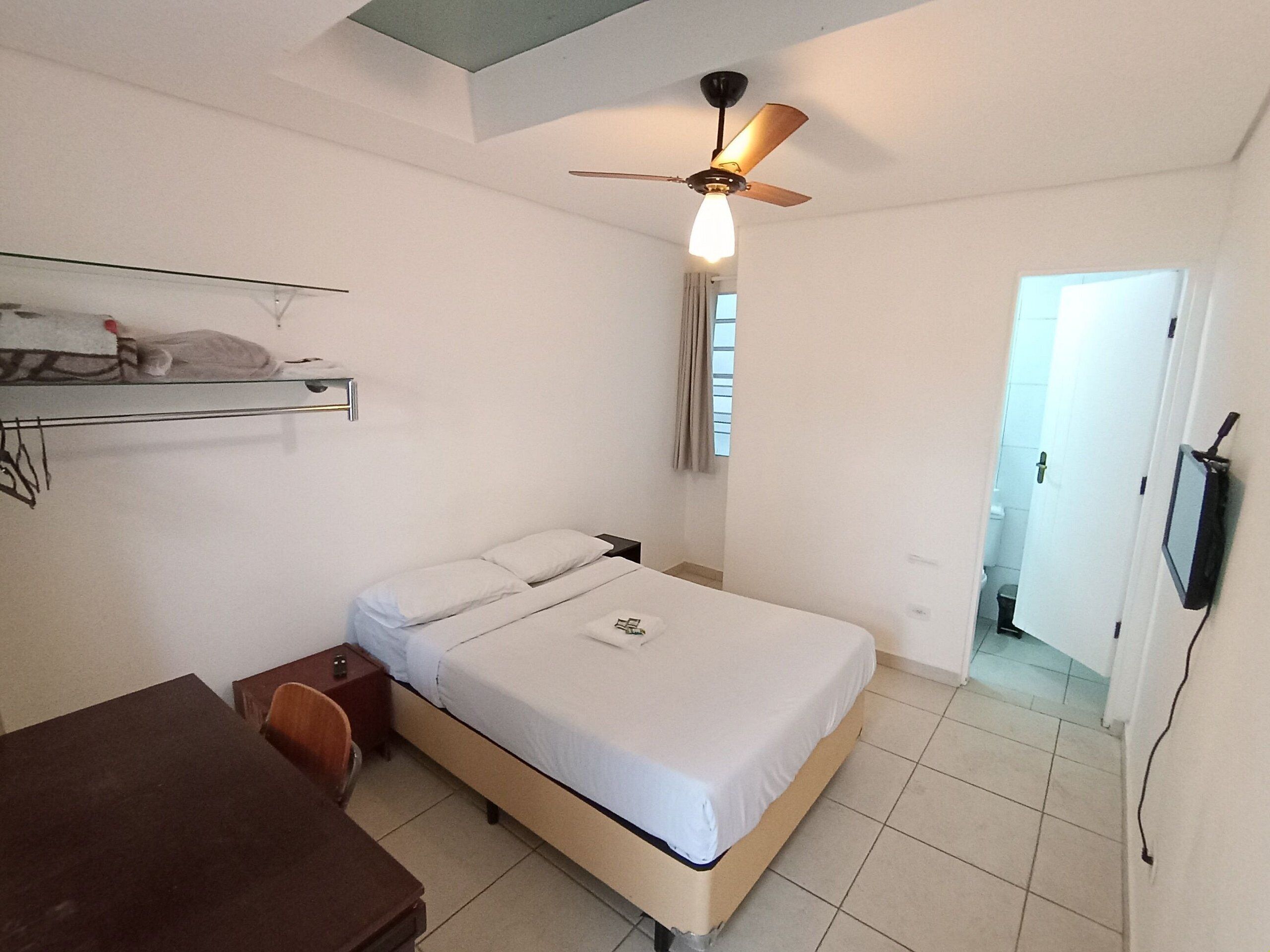 Une chambre à coucher avec un lit, un bureau, une chaise, un ventilateur de plafond et un téléviseur fixé au mur.