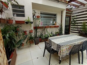 Un patio con una mesa y sillas, rodeado de macetas con plantas y una ventana.