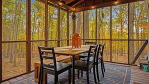 Uma sala de jantar com mesa e cadeiras, janelas amplas e vista para uma floresta.