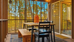 Uma área de jantar com mesa e cadeiras, um banco, um lanterna decorativa e vista para árvores através de grandes janelas.