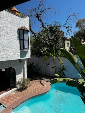 Zona de piscina con un edificio blanco, un camino de ladrillo y abundante vegetación.