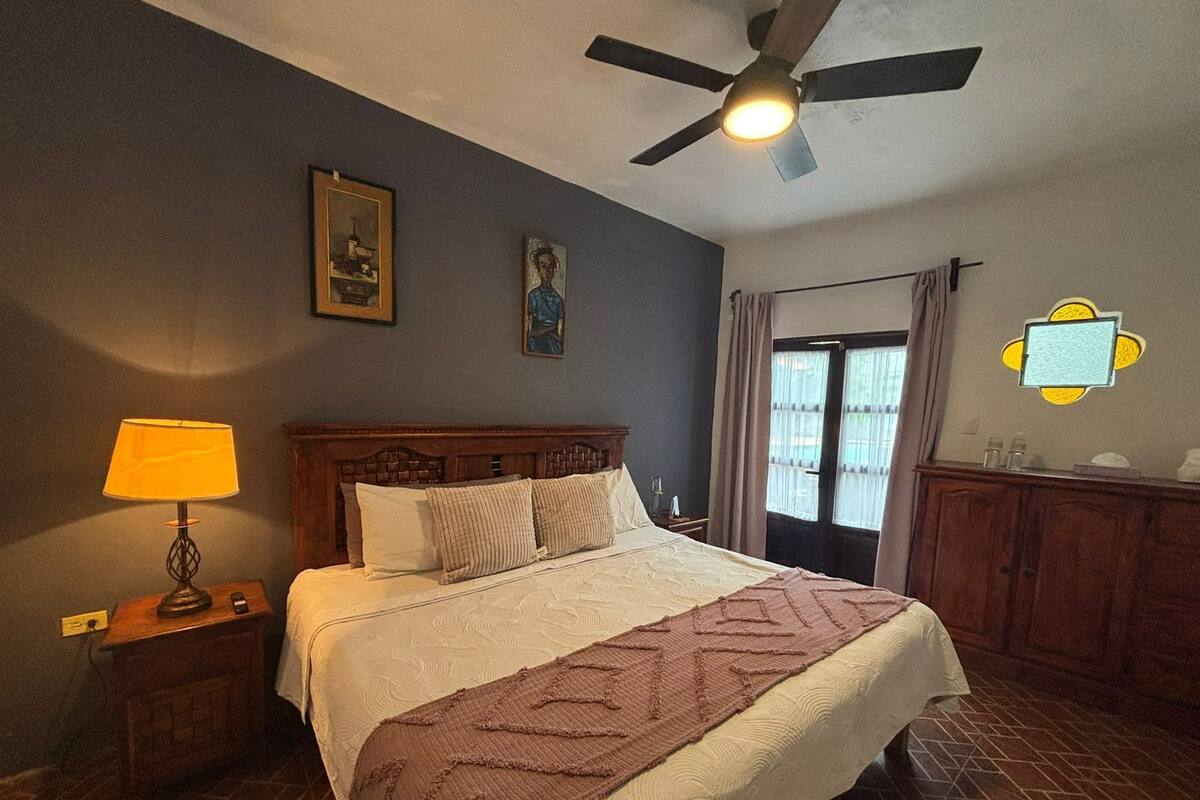 Une chambre à coucher avec un lit, une table de chevet, une lampe, un ventilateur de plafond et une fenêtre avec des rideaux.
