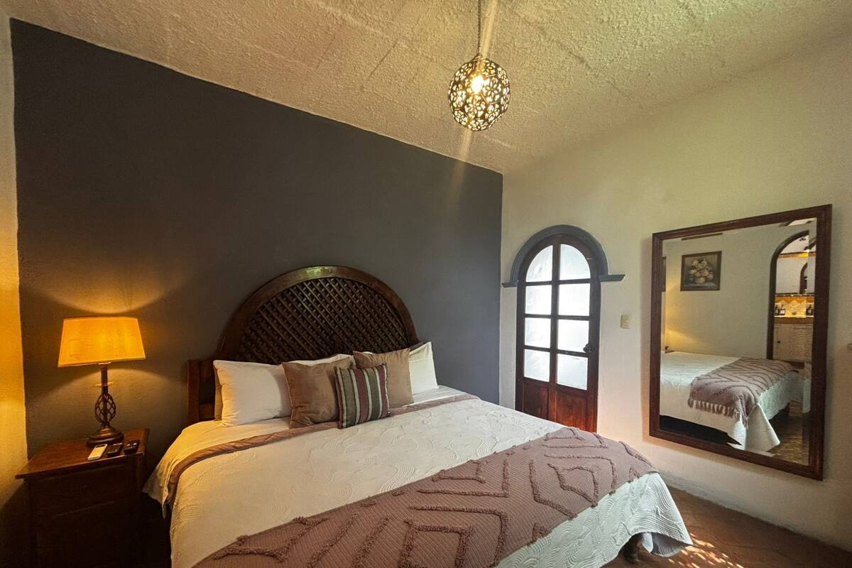 Une chambre à coucher avec un grand lit, une table de chevet, une lampe, un miroir et une fenêtre donnant sur l’intérieur.