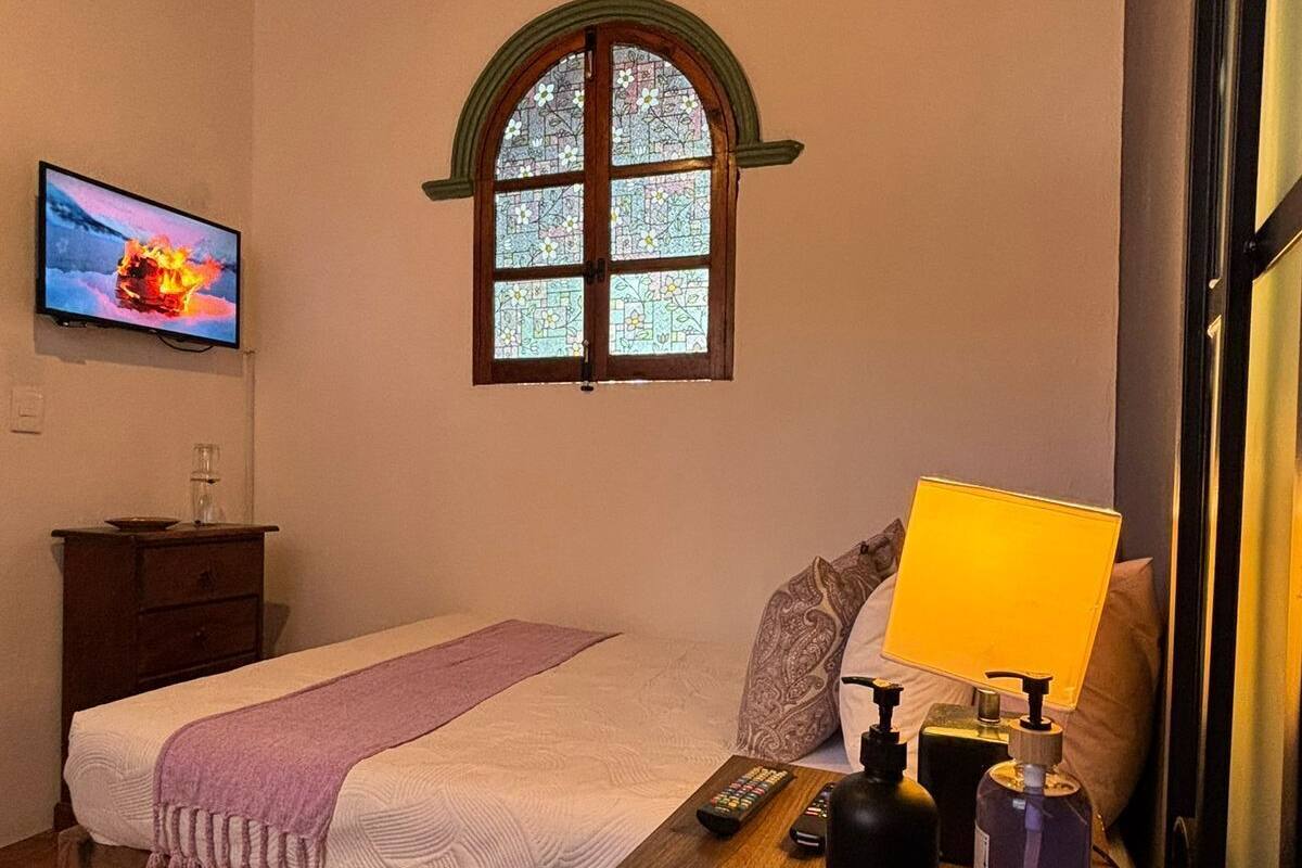 Une chambre à coucher avec un lit, un bureau en bois, une télévision, une fenêtre avec des vitraux et une porte.