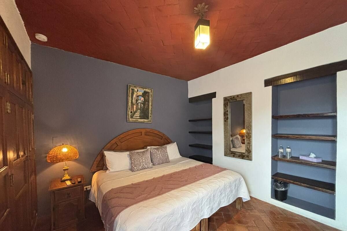 Une chambre avec un lit en bois, une table de chevet avec une lampe, une étagère murale contenant des articles de toilette et un tableau encadré au-dessus du lit.