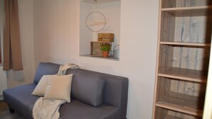 Uma sala de estar moderna com um sofá cinza, uma pequena estante de madeira com uma placa de boas-vindas e uma planta em vaso.