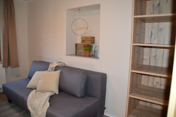 A modern living room with a grey sofa, a small wooden shelf with a welcome sign, and a potted plant.