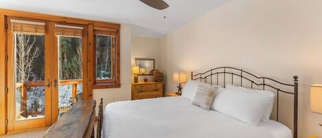 A bedroom with a large bed, wooden headboard, a ceiling fan, and a view of trees through the windows.