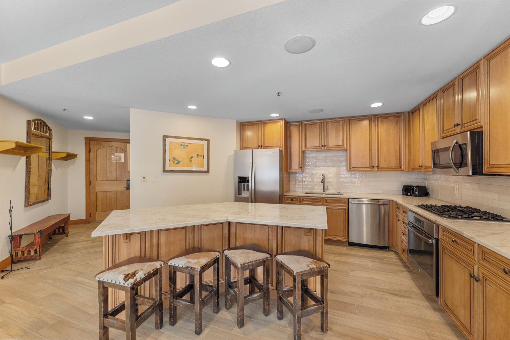 A modern kitchen with wooden cabinets, a large island with stools, stainless steel appliances, and recessed ceiling lights.