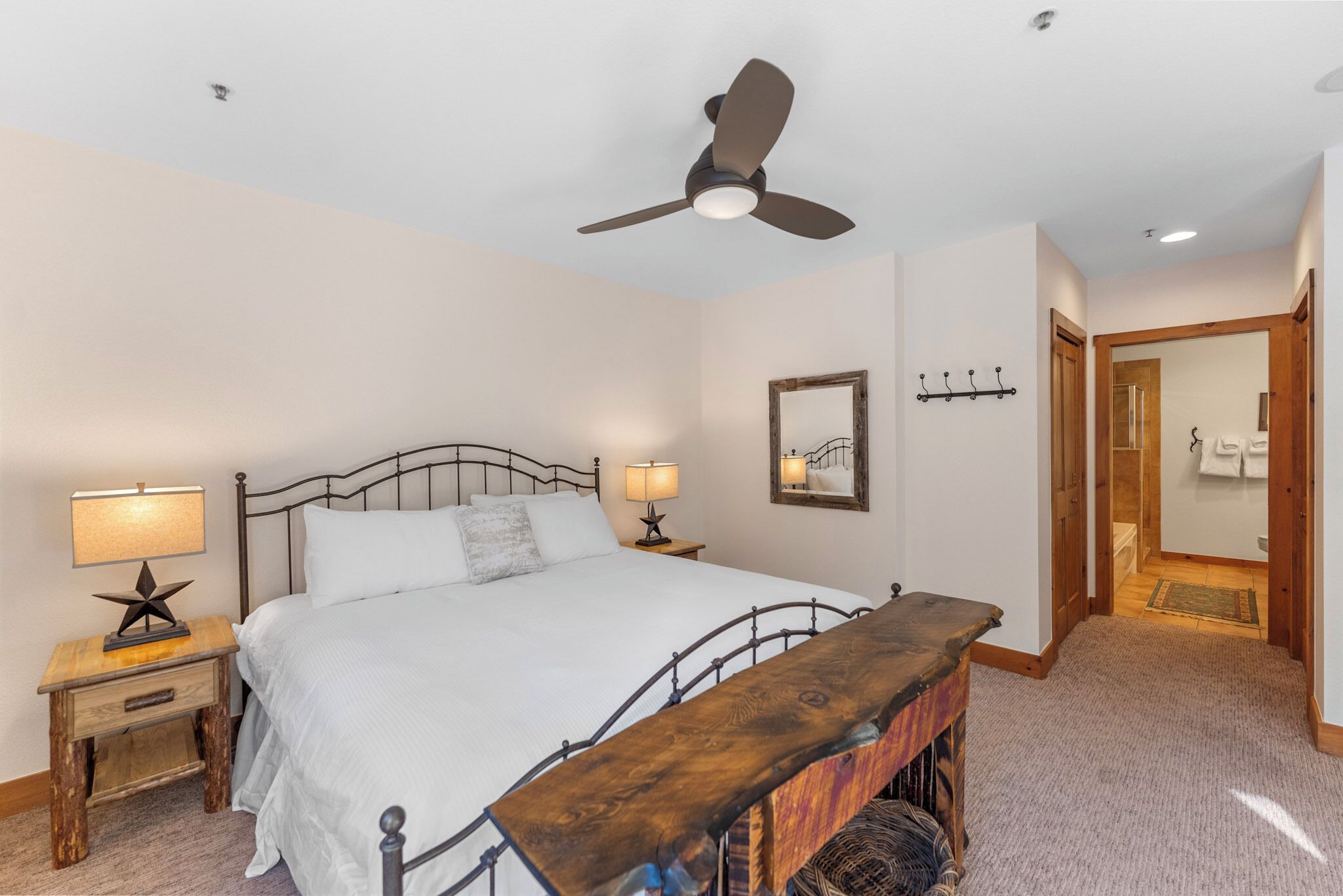 A bedroom with a bed, two bedside tables with lamps, a ceiling fan, a mirror, and a wooden bench.