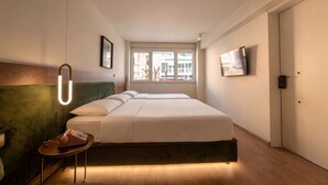 A modern hotel room with a large bed, a small side table, a green headboard, a window with a view, and a TV mounted on the wall. - Pillow by the Sea (Thessaloniki)