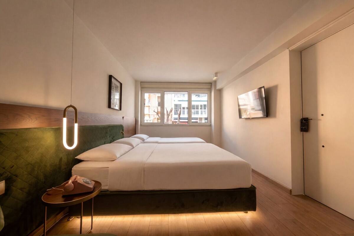 Habitación de hotel moderna con una cama grande, una mesita auxiliar pequeña, un cabecero verde, una ventana con vistas y un televisor montado en la pared.