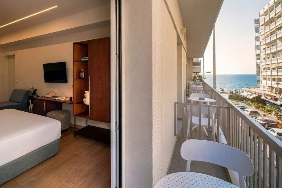 Habitación de hotel con cama, escritorio, silla, televisión y balcón con vista al mar y edificios.