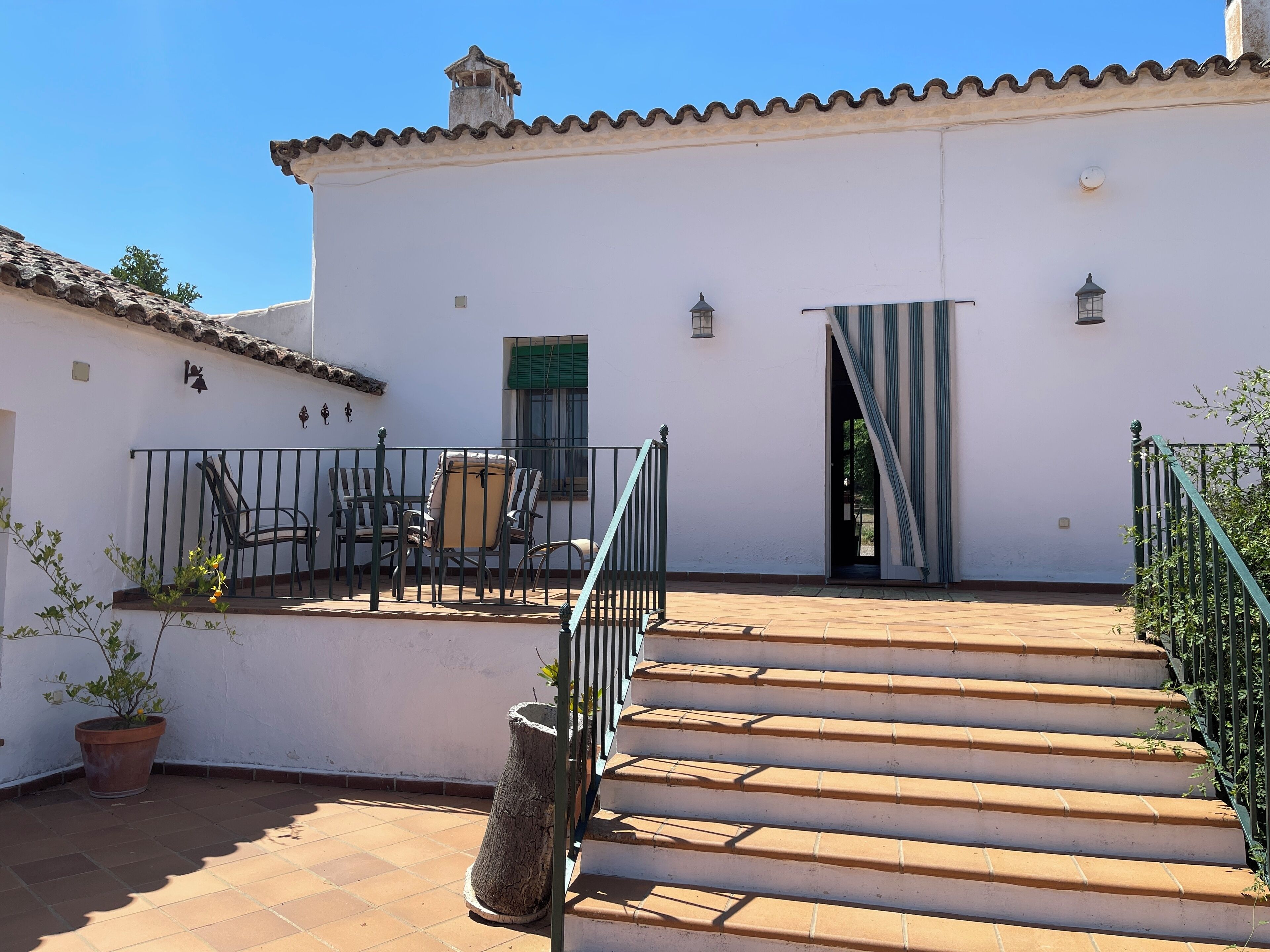 Un edificio blanco con techo de tejas, una escalera que conduce a una puerta y un pequeño patio con sillas y una planta en maceta.