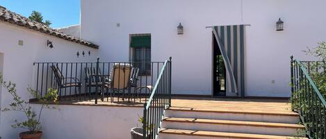 Un edificio blanco con techo de tejas, una escalera que conduce a una puerta y un pequeño patio con sillas y una planta en maceta.