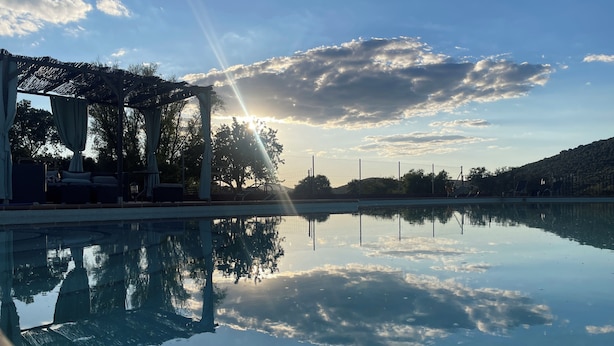 A clear pool with a sun reflecting off the water, surrounded by a covered seating area and trees. - Cortijo Huerta Galindo (Alanís)