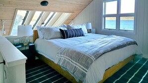 Een slaapkamer met een houten plafond, een groot bed met witte lakens, twee nachtkastjes met lampen en uitzicht op de zee door een raam.