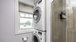 Ein Waschbereich mit gestapelten Waschmaschinen und Trocknern, ein Fenster mit Jalousien und eine geflieste Duschwand.