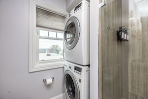 A laundry area with stacked washers and dryers, a window with blinds, and a tiled shower wall.