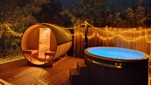 Eine Holztonne als Hot Tub und ein raundes Zimmer aus Holz mit einem Schild mit der Aufschrift 'HOME DELUXE', vor einer Kulisse aus Bäumen und Lichterketten.