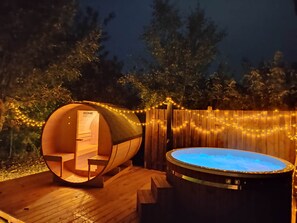 A wooden hot tub and a wooden barrel-shaped room with a sign reading 'HOME DELUXE' against a backdrop of trees and string lights.