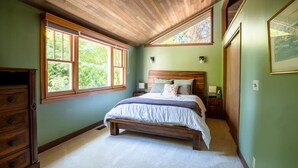 Een slaapkamer met een houten bed, een dressoir en een groot raam met uitzicht op groen.