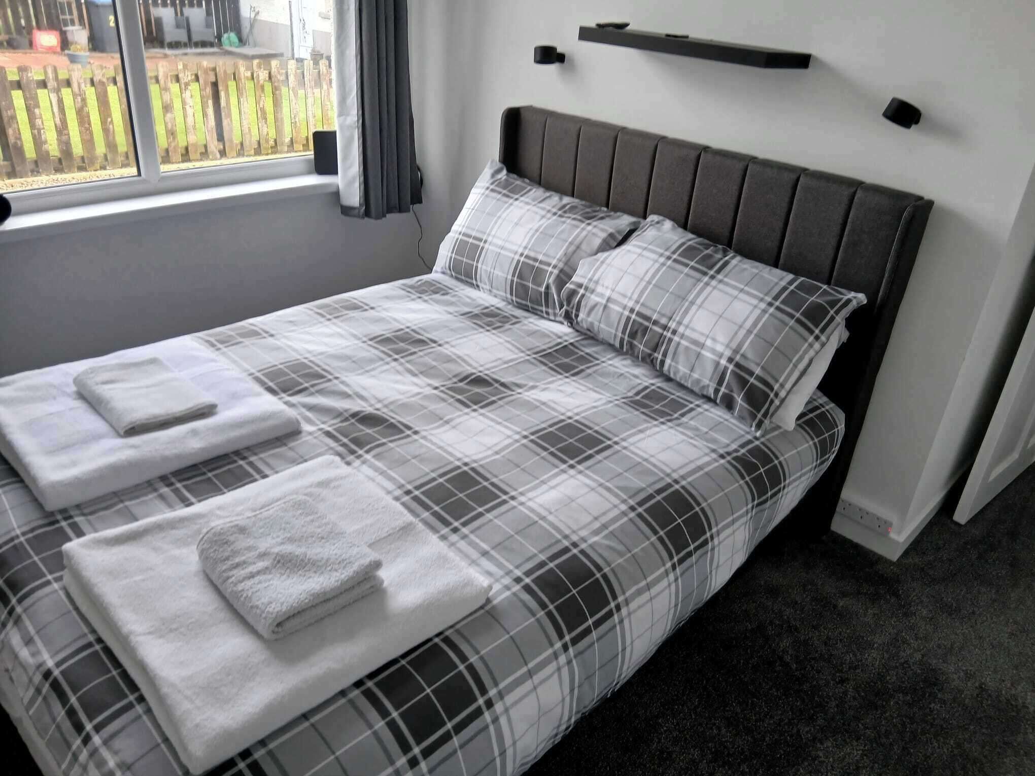 A neatly made bed with a plaid bedspread and pillows, a bedside table with a lamp, and a window with curtains.