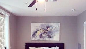 A bedroom with a bed, a ceiling fan, a wall-mounted abstract painting, and a bedside table with a lamp.