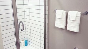 A bathroom with a glass shower door, white tiled walls, a wooden floor, and a towel rack with two white towels.