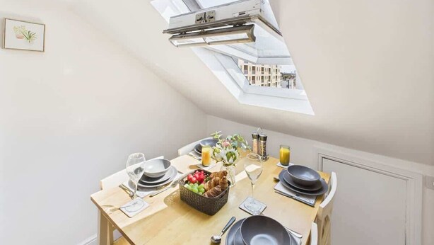 A dining area with a table set for two, a window with a view of buildings, and a skylight above the table.