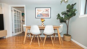 Un área de comedor con una mesa de madera y cuatro sillas blancas, un cuadro enmarcado en la pared y una planta en maceta.