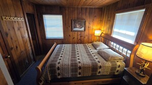A wooden room with a bed, two windows with blinds, a framed picture on the wall, and a lamp on a bedside table. - Cabin 2 @ Lazy Day Cabins (Mayhill)