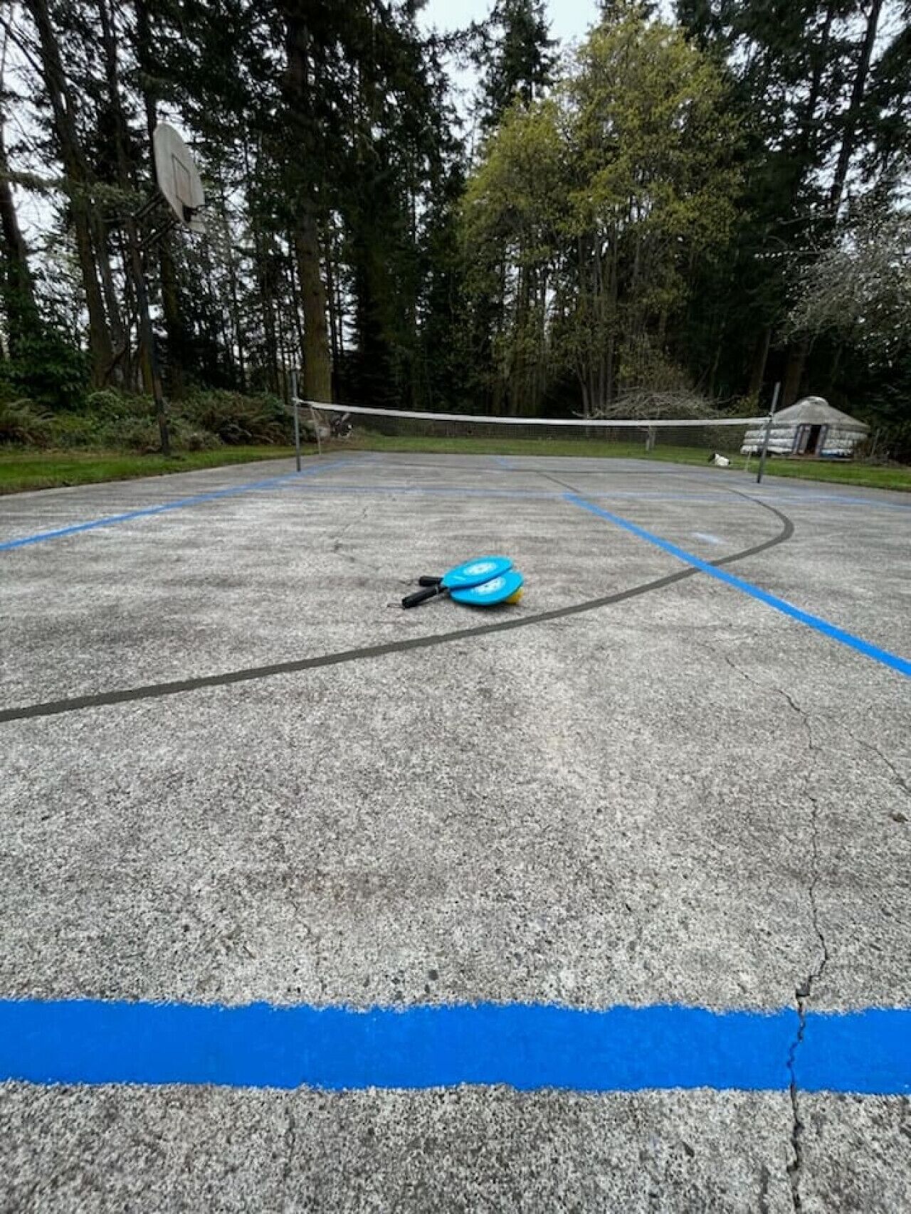 Uma quadra esportiva de concreto com linhas azuis, cercada por árvores e uma pequena construção ao fundo.