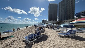A beach with lounge chairs and umbrellas, a clear sky, and tall buildings in the background. - Luxury 3-BR Apartment with Stunning City (Hallandale Beach)