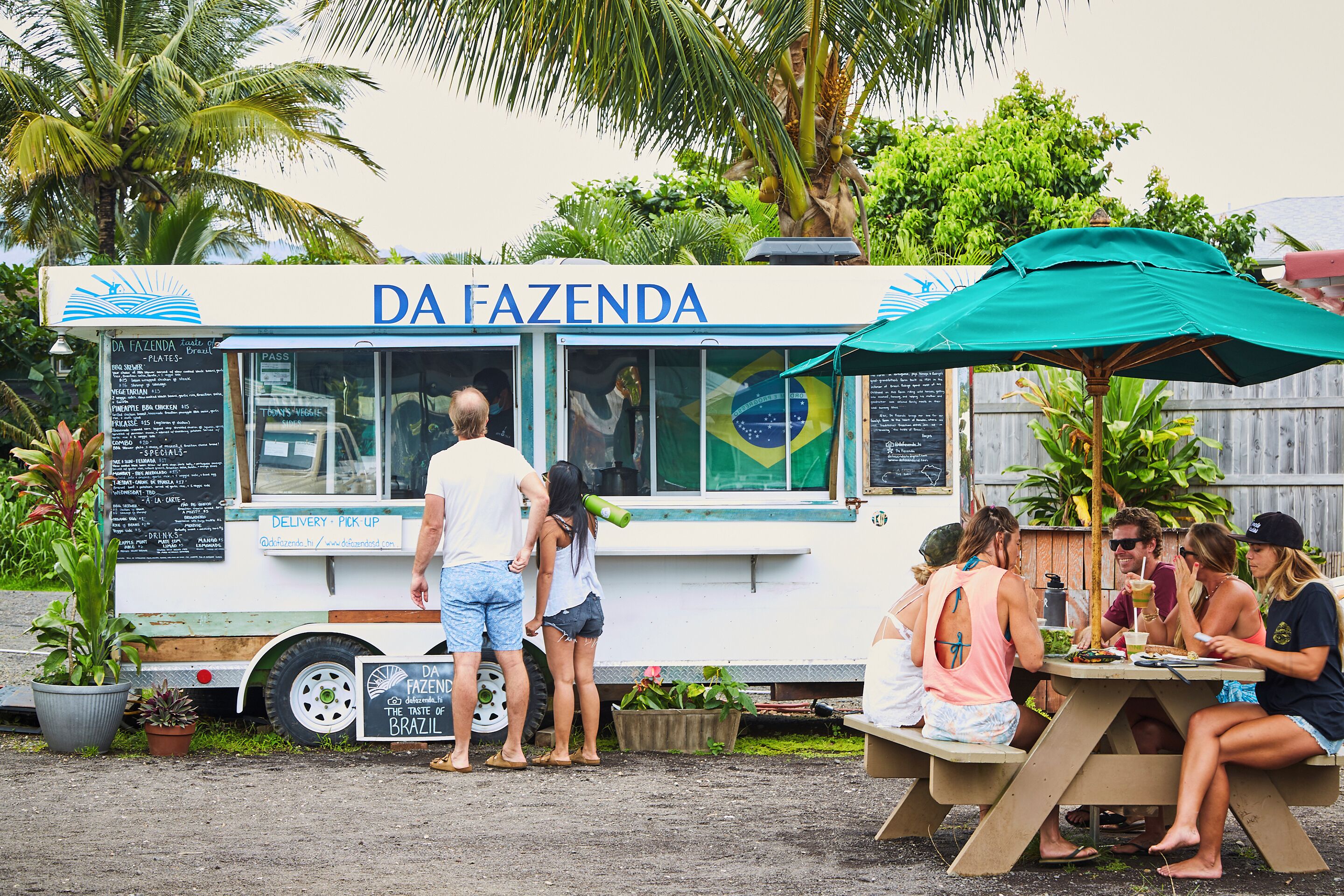 Un food truck anomenat DA FAZENDA amb un quadre de menú, un grup de persones dempeus i assegudes al seu voltant, i una paraigua verda que ofereix ombra.