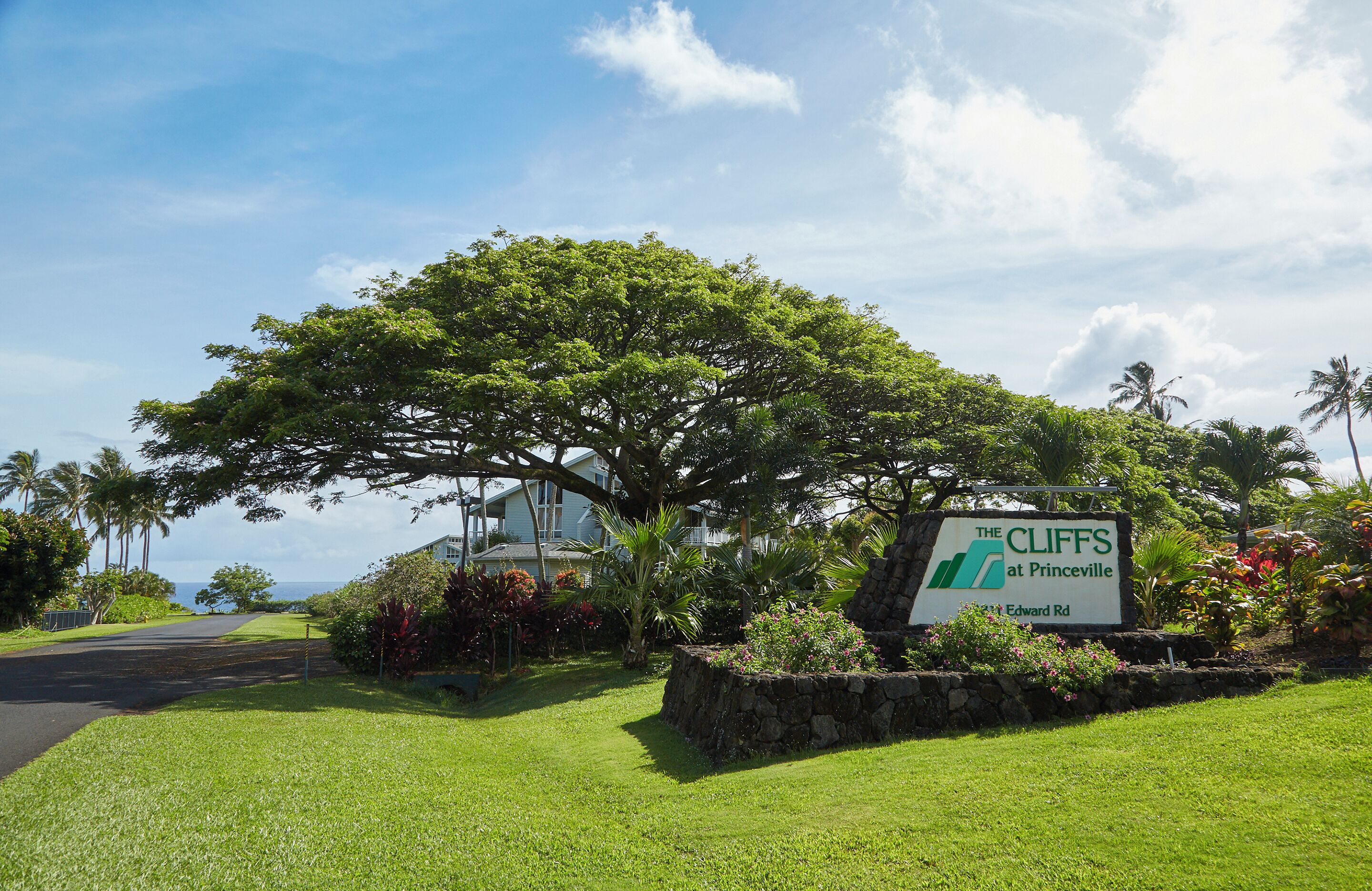 A sign for 'The Cliffs at Princeville' with a view of a lush green landscape and a large tree.