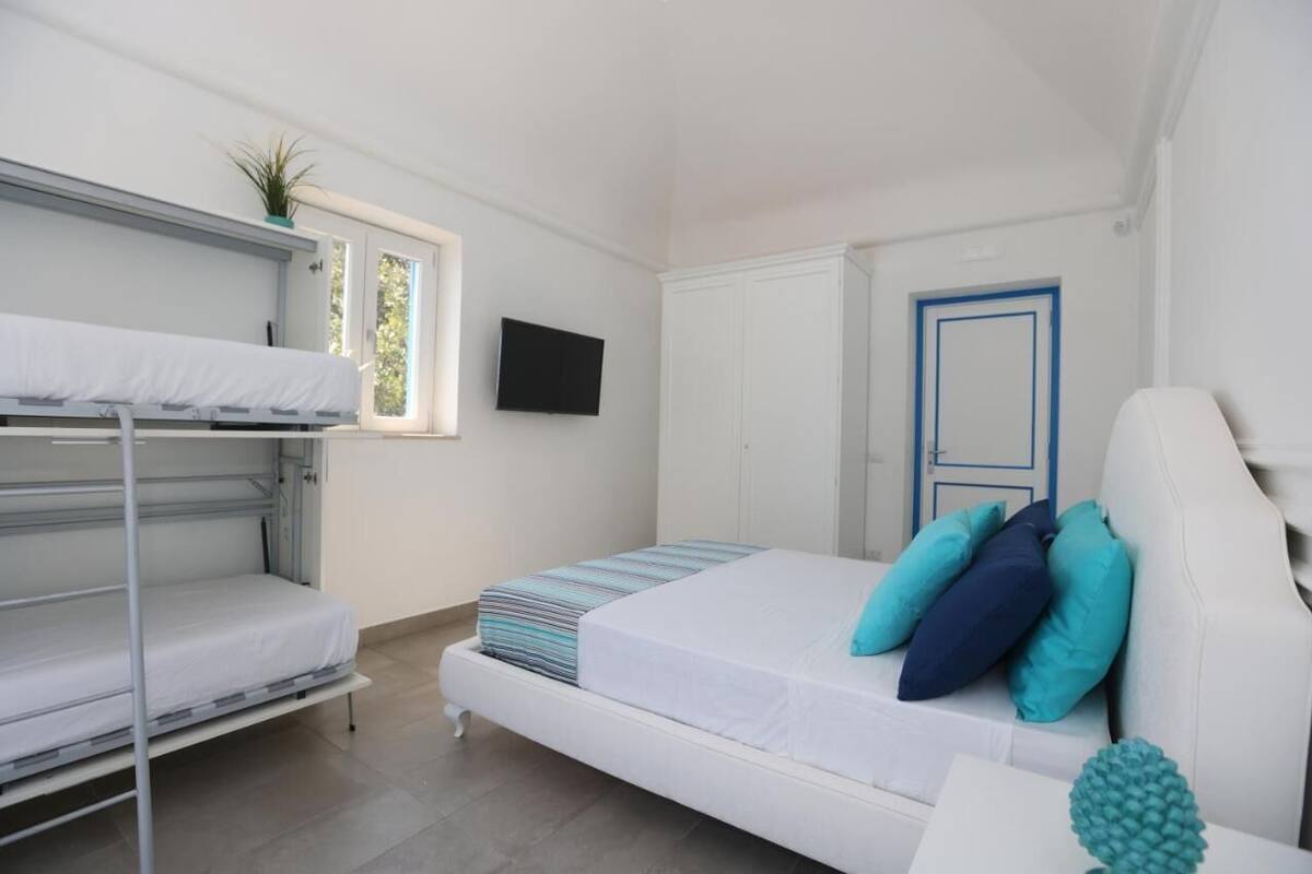 A bedroom with a bunk bed, a single bed, a TV mounted on the wall, and a window with a view of greenery.