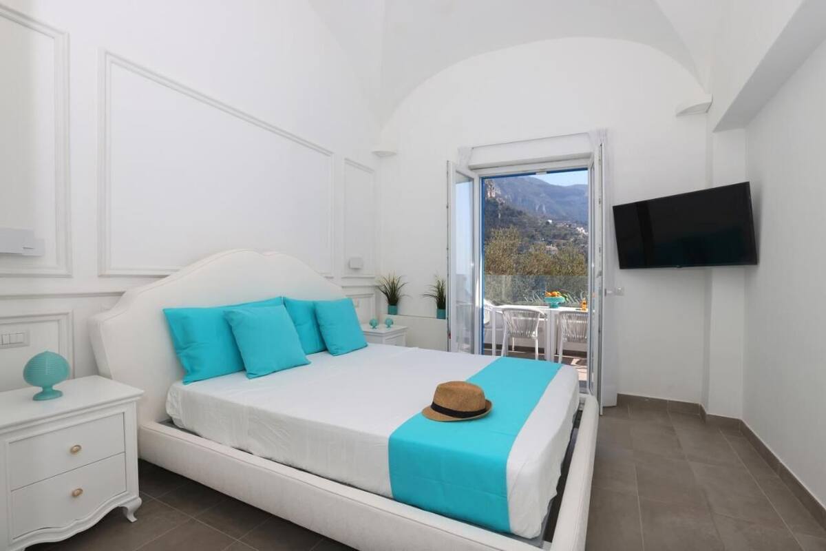 A bedroom with a bed, a nightstand, a TV mounted on the wall, and a balcony with a view of the mountains.