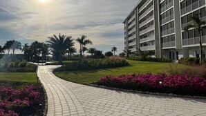 A pathway leading to a modern building with multiple balconies, surrounded by well-maintained greenery and flowering plants. - Oceanfront Condo at Bonita Beach Club – Pool, Tennis, Private Beach Access! (Bonita Springs)