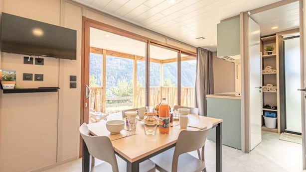 Een eetgedeelte met een tafel gedekt voor twee, een keuken met een koelkast, en een schuifdeur naar een balkon met uitzicht op de bergen.