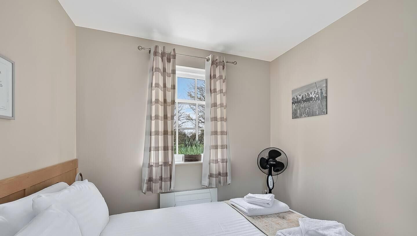 A bedroom with a bed, a window with curtains, a fan, and a wall-mounted picture.
