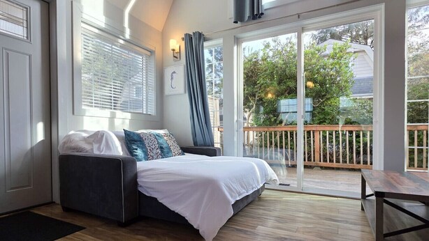 Ein gemütliches Zimmer mit einem Bett, einem Fenster mit Jalousien, einem Balkon mit Holzterrasse und Blick ins Grüne.