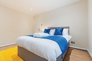 Een opgemaakt bed met wit- en blauw beddengoed, een grijs hoofdbord en een geel vloerkleed.