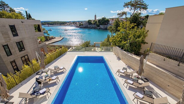 A rectangular pool with lounge chairs and umbrellas, overlooking a body of water with boats and a distant town. - HADRIE Luxury Resort and Villas Krk (Krk)