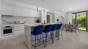 A modern kitchen with white cabinets, a marble countertop, and a built-in oven. There are blue bar stools and a dining area with a table and chairs. - HADRIE Luxury Resort and Villas Krk (Krk)