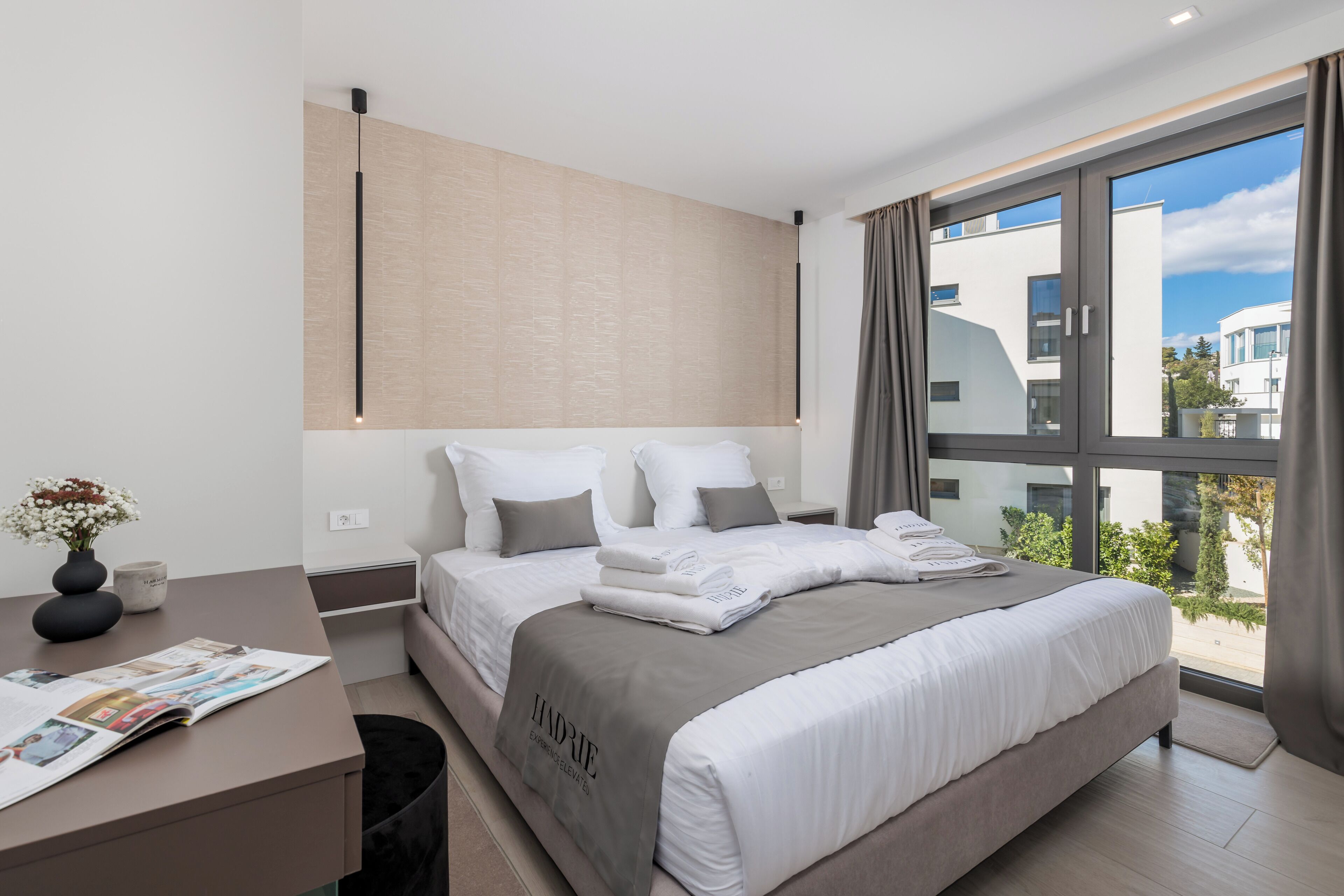 Ein modernes Hotelzimmer mit einem großen Bett, einem Schreibtisch mit einer Zeitschrift und Blick auf die Stadt durch große Fenster.