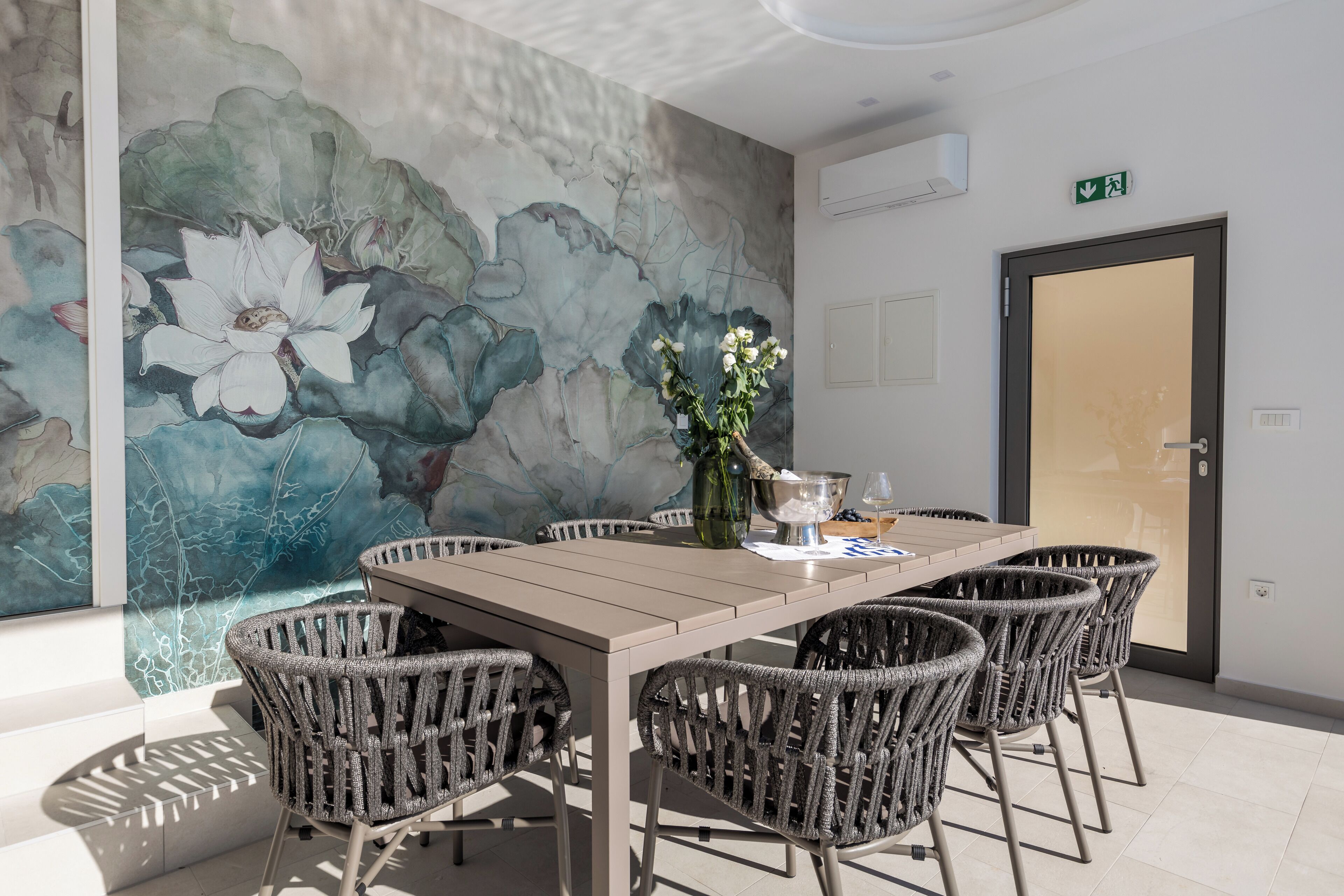 Un comedor con una mesa y sillas de mimbre, un mural en la pared y una puerta con un letrero de salida.