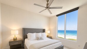 Quarto com cama, mesas de cabeceira, ventilador de teto e uma grande janela com vista para a praia.