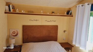 Una camera da letto con una testiera marrone, una lampada da comodino, uno smartphone Galaxy S21 5G sul tavolino e uno scaffale con libri e oggetti decorativi.